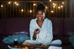 A woman sitting on a couch at night, wearing cozy loungewear and holding a remote control, smiling as she watches TV. Soft string lights are glowing in the background, creating a warm and relaxed atmosphere. The Best African Movies on Netflix