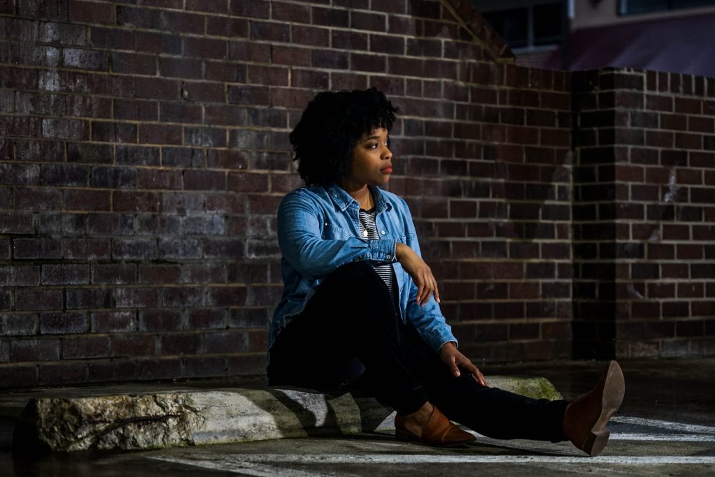 A thoughtful woman sitting outdoors against a brick wall, reflecting on the challenges and opportunities of moving abroad and fitting in. Featured on Raided by Tintin.