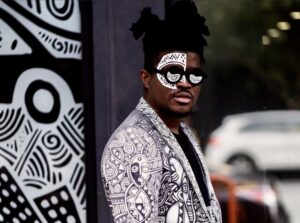 Portrait of Laolu Senbanjo wearing his signature Sacred Art design, with his intricate artwork as a backdrop. Featured on Raided by Tintin.