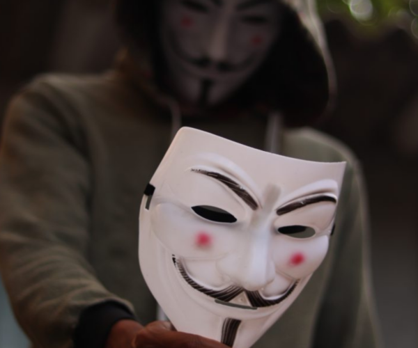 A person in a hoodie holding a Guy Fawkes mask, symbolizing the deceptive tactics of impersonators who mimic others' identities online. Featured in the article 'Types of Cyberbullies: Tackling Digital Harassment with Confidence' on Raided by Tintin.