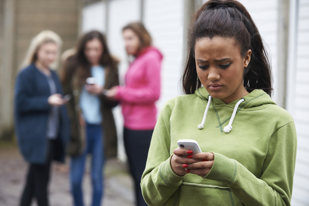 Teen girl looking distressed while checking her phone, with a group of peers in the background, representing the impact of different types of cyberbullies. Featured in the article 'Types of Cyberbullies: Tackling Digital Harassment with Confidence' on Raided by Tintin.