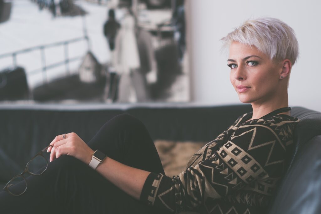 Confident woman with short, stylish early grey hair, sitting on a sofa in a patterned sweater, embodying grace and elegance. Perfect representation for the article 'Silver Secrets: Embracing Early Grey Hair with Style' by Raided by Tintin.