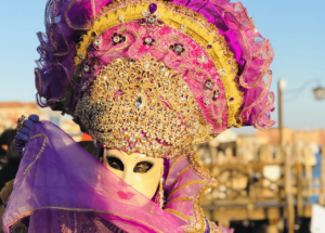 A masked participant adorned in a vibrant purple and gold costume at the Venice Carnival, showcasing the elegance of traditional festivals. Featured in the article 'Coolest Traditional Festivals: Celebrating Culture in Style' on Raided by Tintin.