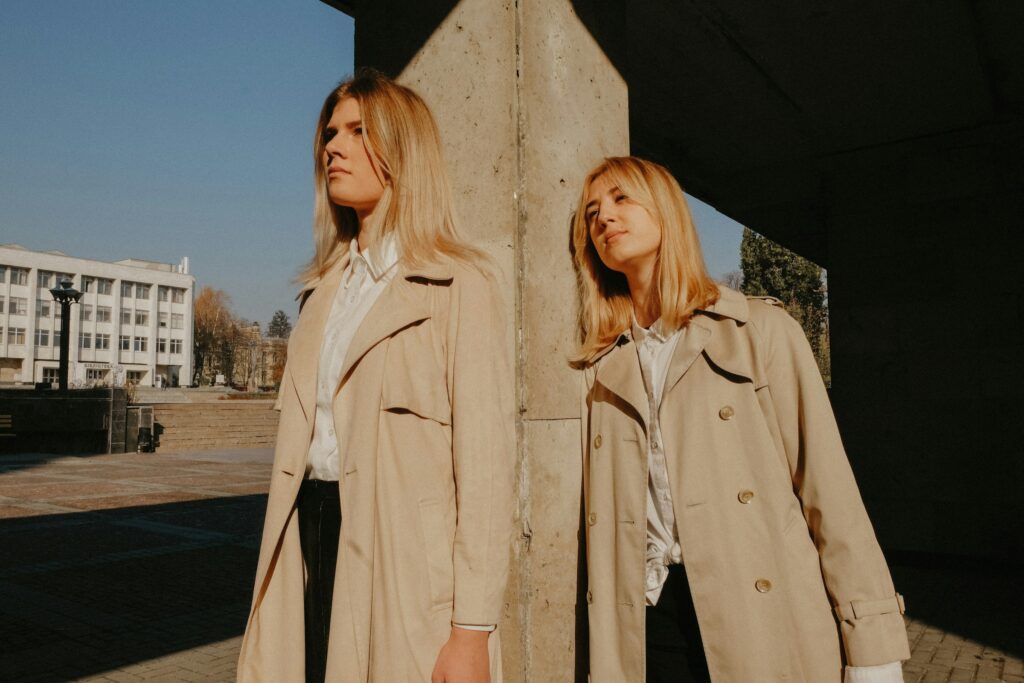 Two women standing confidently in beige trench coats under sunlight, blending elegance and casual style. Photo by Masha Kotliarenko on Unsplash.