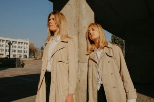 Two women standing confidently in beige trench coats under sunlight, blending elegance and casual style. Photo by Masha Kotliarenko on Unsplash.